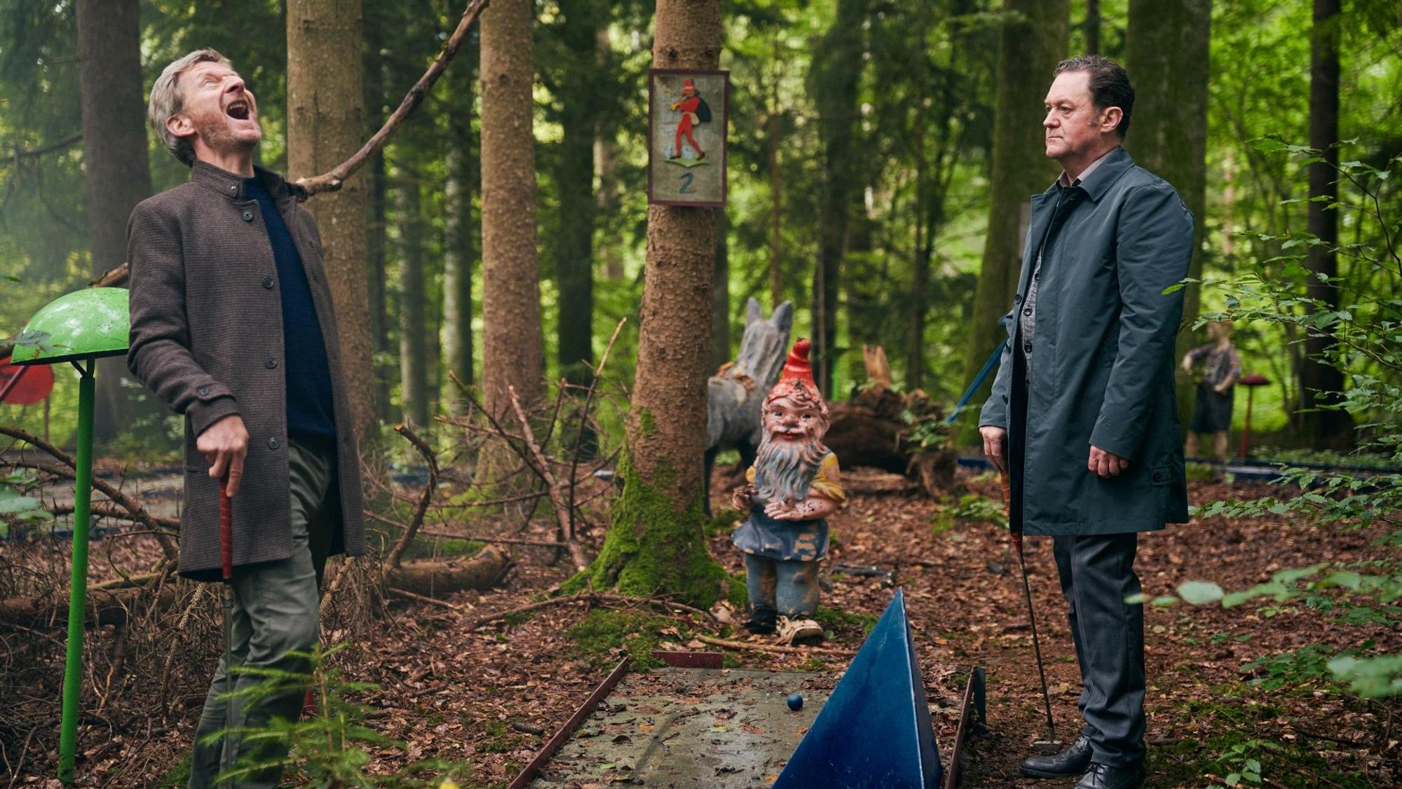 Zwei Männer stehen im Wald, einer lacht lauthals.