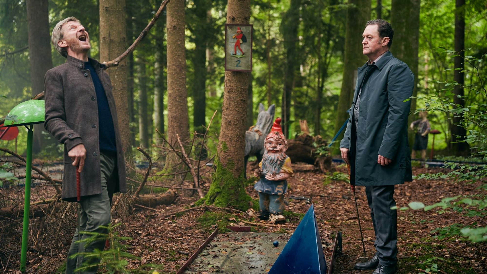 Zwei Männer stehen im Wald, einer lacht lauthals.