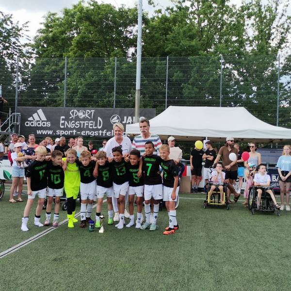 Eine Fußballmannschaft, bestehend aus Jungs, stehen in einer Reihe. Im Hintergrund ist Handballlegende Heiner Brand zu sehen.
