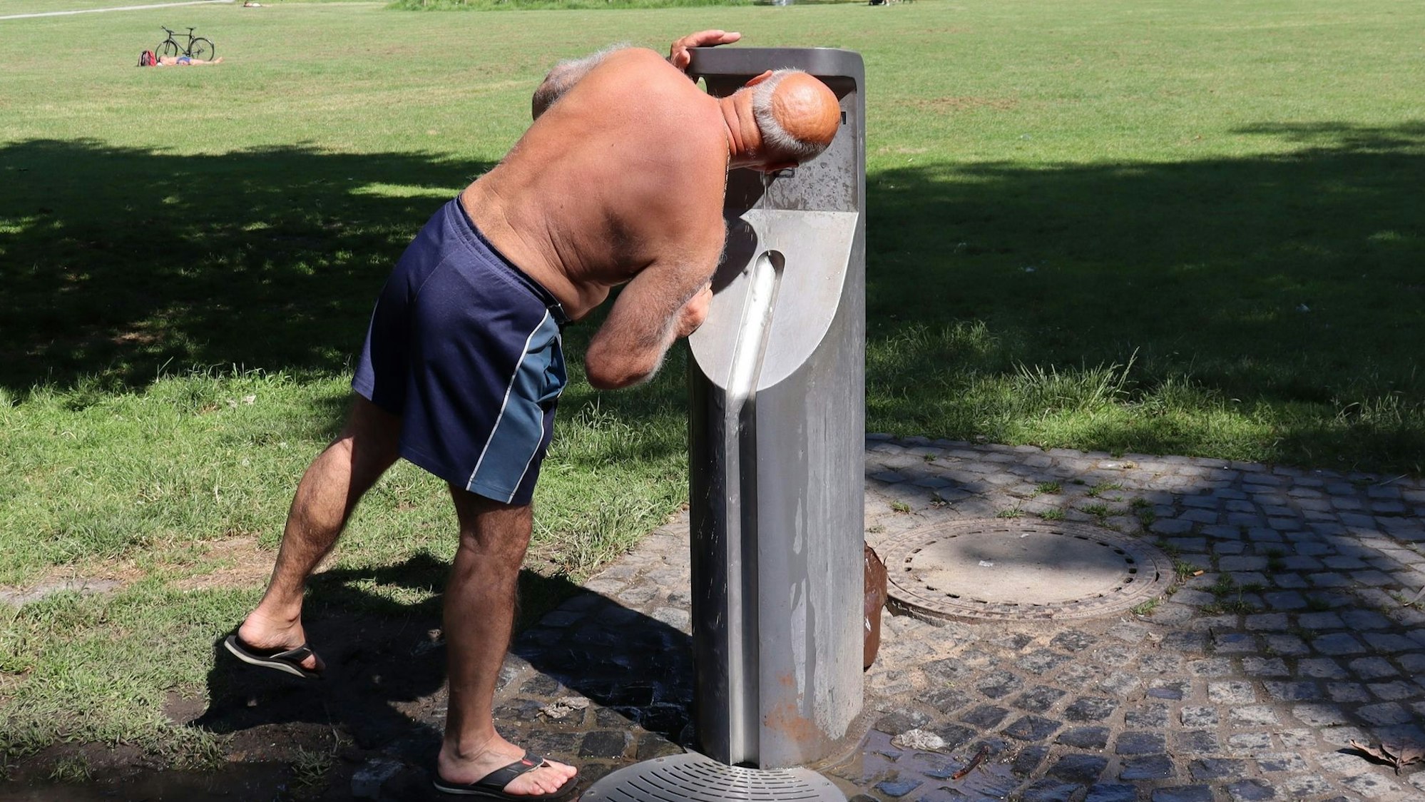 Ein Mann trinkt im Park aus einem Trinkwasserspender.