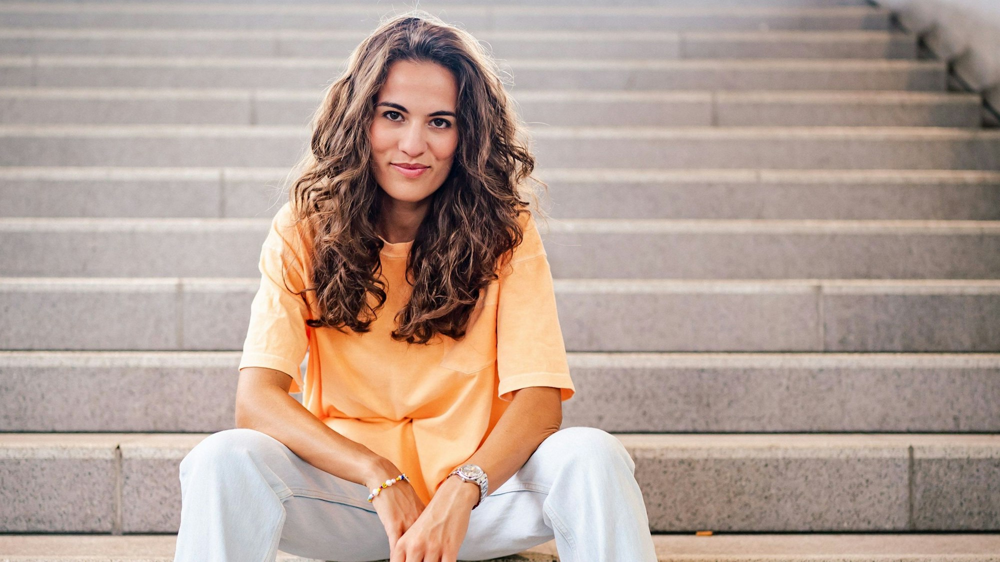 Mona Ameziane sitzt auf einer sterinernen Treppe. Sie hat die Arme auf die Oberschenkel gelehnt und trägt ein oranges Top.