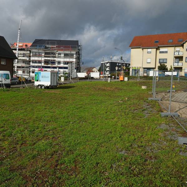 Rechts am Bildrand sind die Absperrgitter um eine grüne Wiese zu sehen. Im Hintergrund sowie am linken Bildrand sind Wohnhäuser zu sehen.