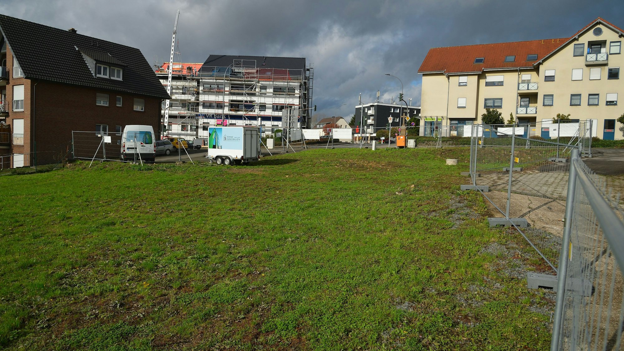 Rechts am Bildrand sind die Absperrgitter um eine grüne Wiese zu sehen. Im Hintergrund sowie am linken Bildrand sind Wohnhäuser zu sehen.