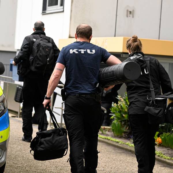 Mitarbeiter der Spurensicherung vor dem Hochhaus, in dem eine Explosion mehrere Einsatzkräfte von Polizei und Feuerwehr schwer verletzt hatte. Ein Streifenwagen ist zu sehen, der Eingangsbereich des Hauses sowie zwei Männer und eine Frau von hinten, wie sie Richtung Eingang gehen.
