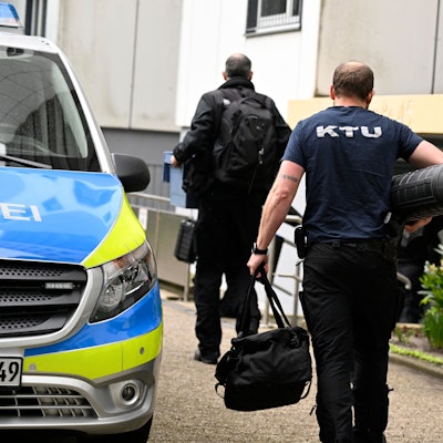 Mitarbeiter der Spurensicherung vor dem Hochhaus, in dem eine Explosion mehrere Einsatzkräfte von Polizei und Feuerwehr schwer verletzt hatte. Ein Streifenwagen ist zu sehen, der Eingangsbereich des Hauses sowie zwei Männer und eine Frau von hinten, wie sie Richtung Eingang gehen.