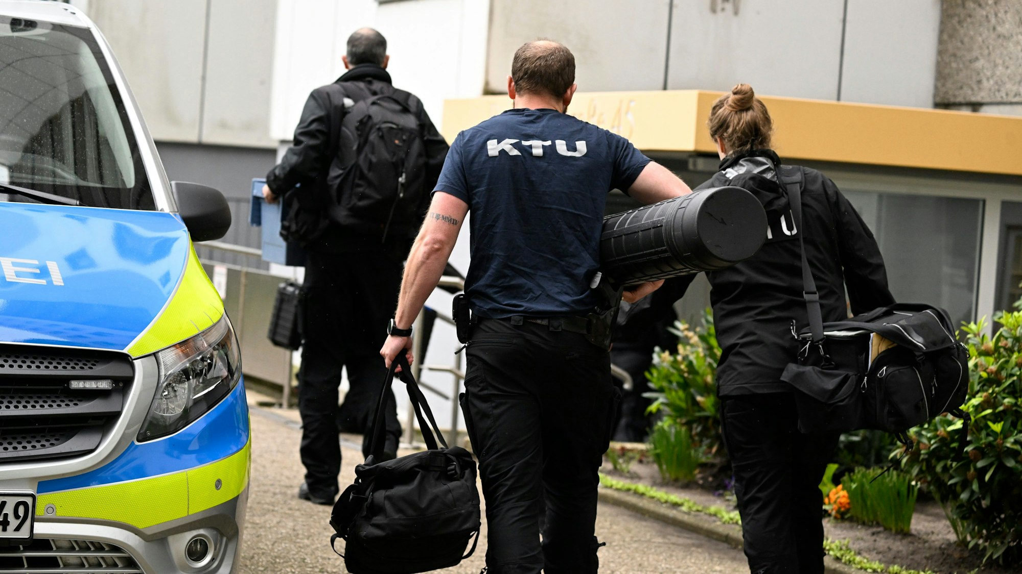 Mitarbeiter der Spurensicherung vor dem Hochhaus, in dem eine Explosion mehrere Einsatzkräfte von Polizei und Feuerwehr schwer verletzt hatte. Ein Streifenwagen ist zu sehen, der Eingangsbereich des Hauses sowie zwei Männer und eine Frau von hinten, wie sie Richtung Eingang gehen.