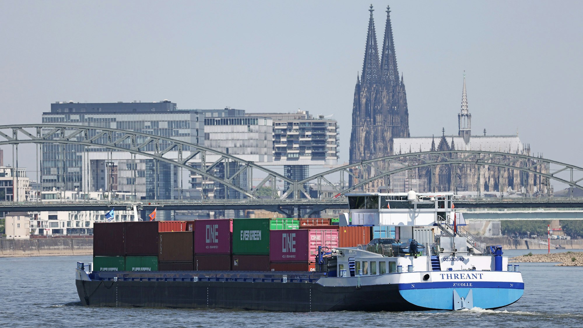 Ein Binnenschiff, mit Containern beladen, fährt in Köln vor dem Dom und den Kranhäusern über den Rhein.