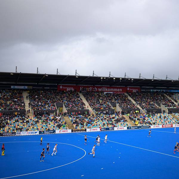 Ein blaues Hokey-Spielfeld ist zu sehen mit laufendem Match. Die Besuchertibüne ist gut besetzt.
