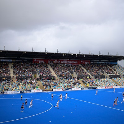 Ein blaues Hokey-Spielfeld ist zu sehen mit laufendem Match. Die Besuchertibüne ist gut besetzt.