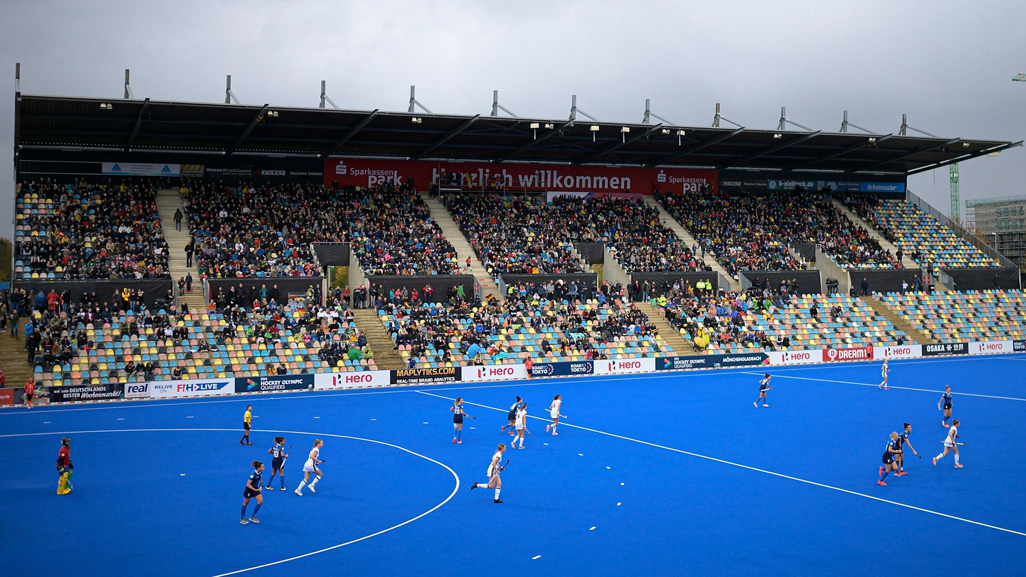 Ein blaues Hokey-Spielfeld ist zu sehen mit laufendem Match. Die Besuchertibüne ist gut besetzt.