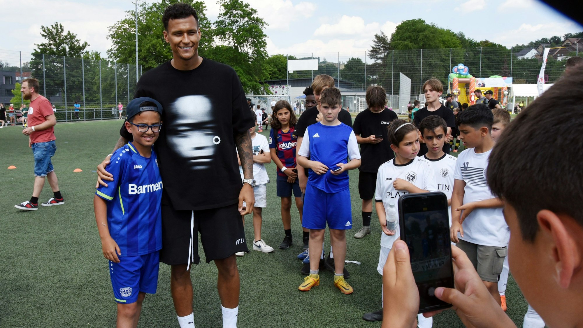 Ein Junge in blauem Trikot lässt sich neben einem Fußball-Profi auf einem Fußballplatz von einem weiteren Jungen mit dem Handy fotografieren.