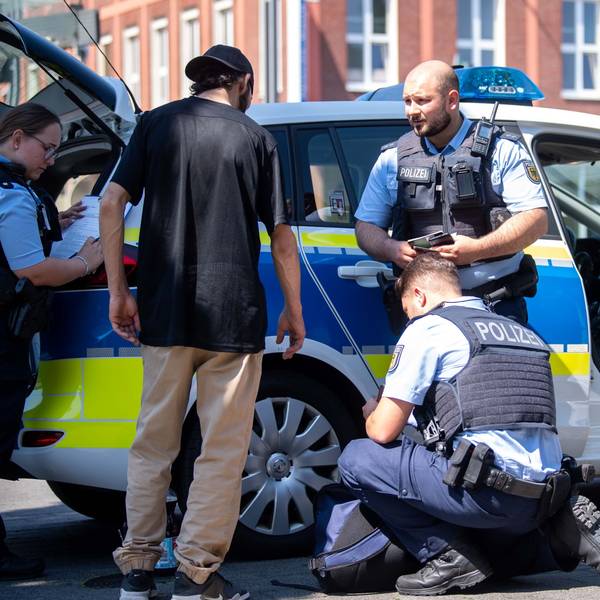 Ein Passant steht vor einem Polizeiwagen. Drei Beamte kontrollieren ihn und nehmen seine Personalien auf.