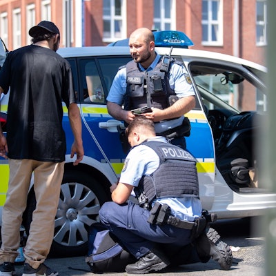 Ein Passant steht vor einem Polizeiwagen. Drei Beamte kontrollieren ihn und nehmen seine Personalien auf.