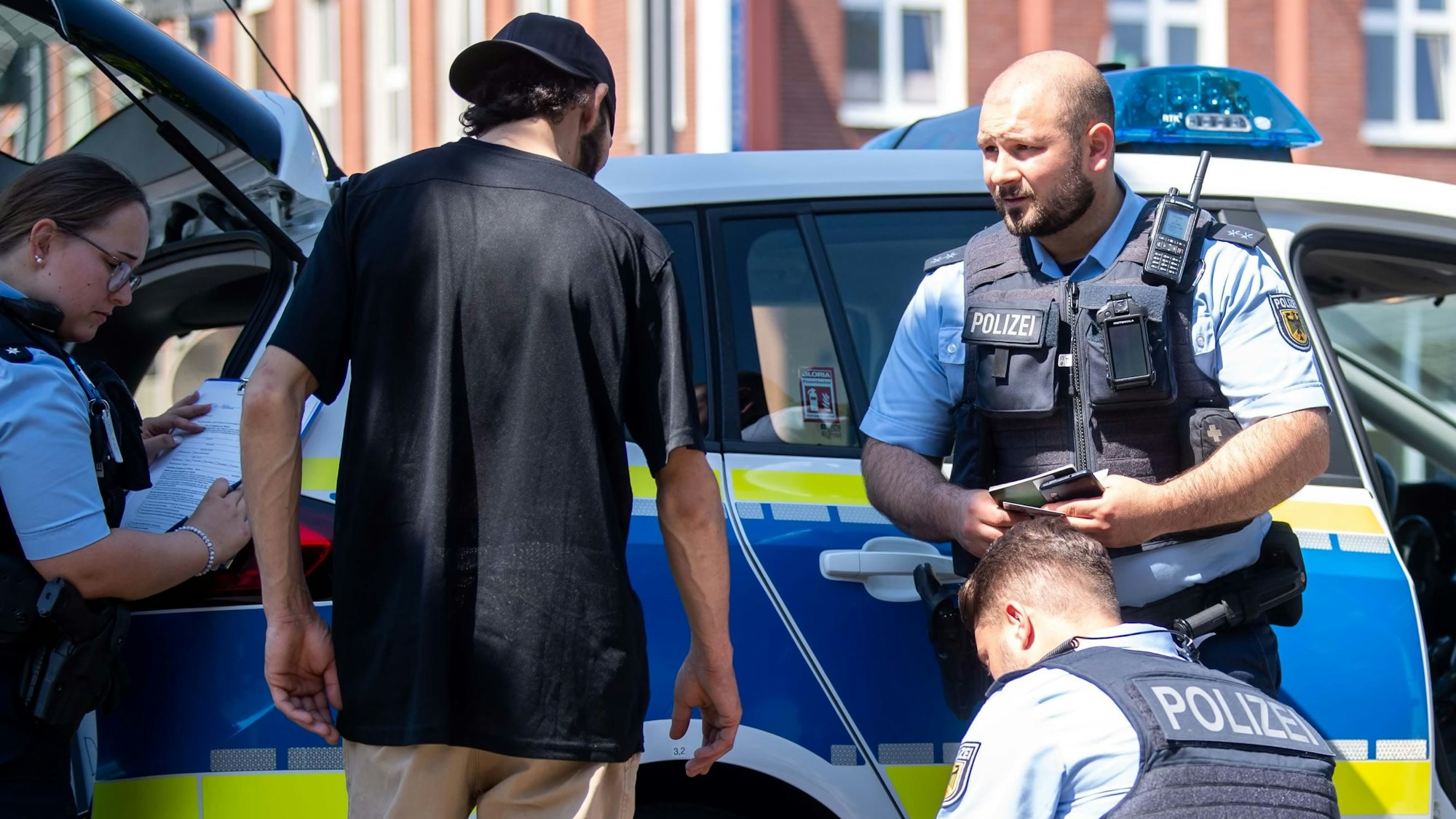 Ein Passant steht vor einem Polizeiwagen. Drei Beamte kontrollieren ihn und nehmen seine Personalien auf.