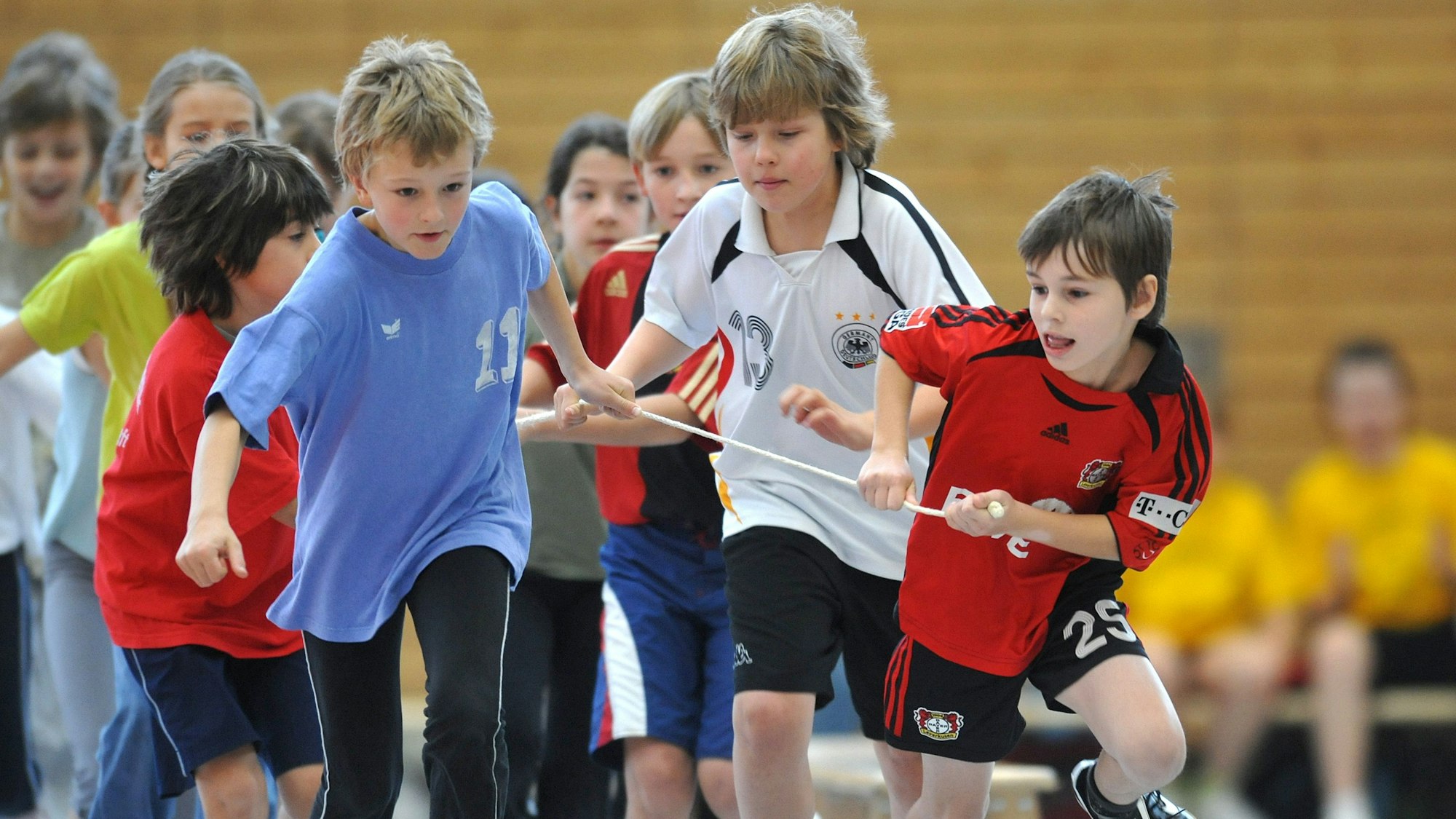 Eine Gruppe von etwa zehnjährigen Kindern übt die Staffelübergabe beim Staffellauf. Paare von je zwei Kindern sind über einen Stab miteinander verbunden. Im Bildhintergrund sitzen Kinder auf einer Bank.