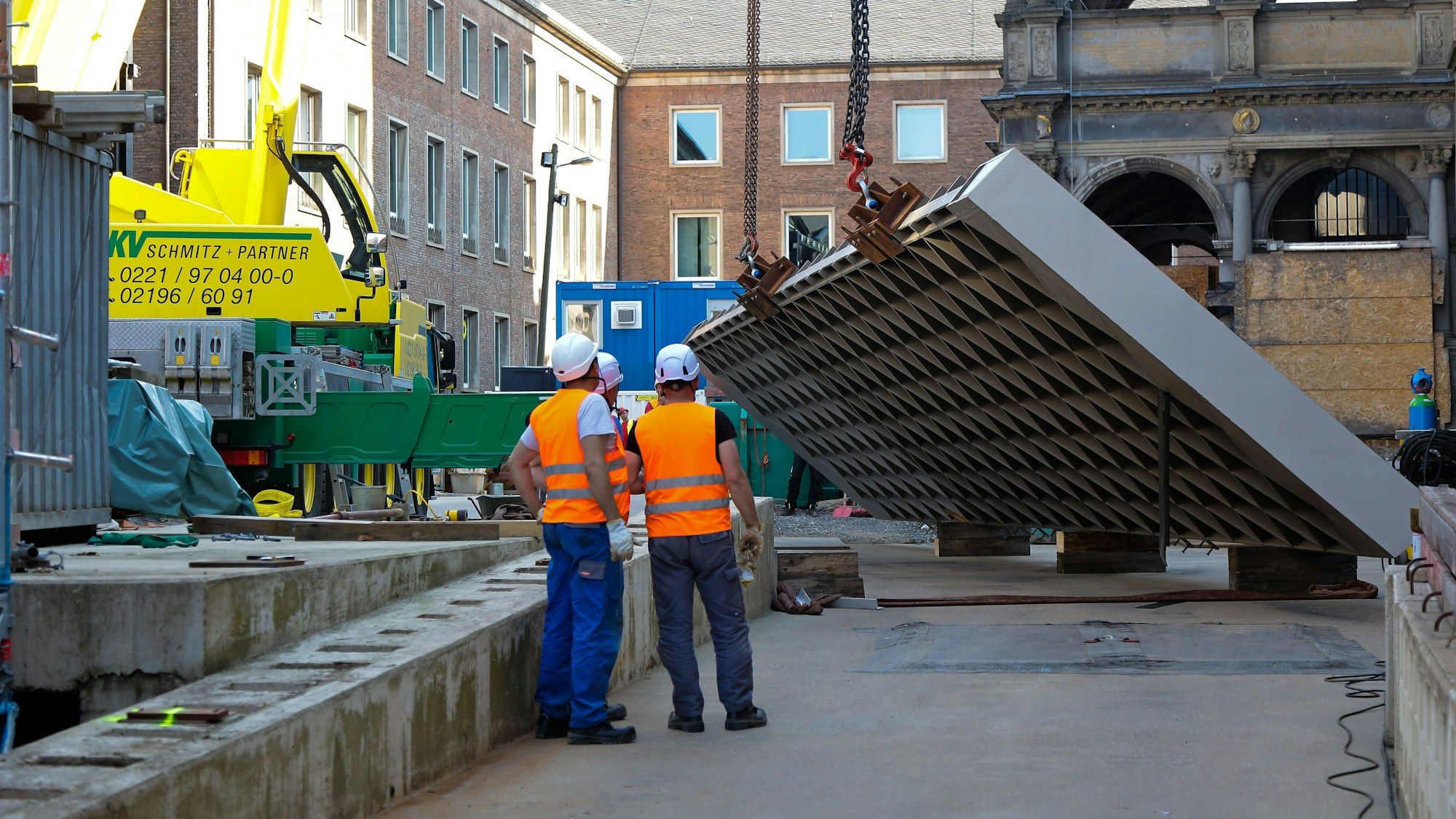 Drei Männer auf der Baustelle der „Miqua“ mit dem fehlendem Seitenteil, welches an einem Kran hängt.