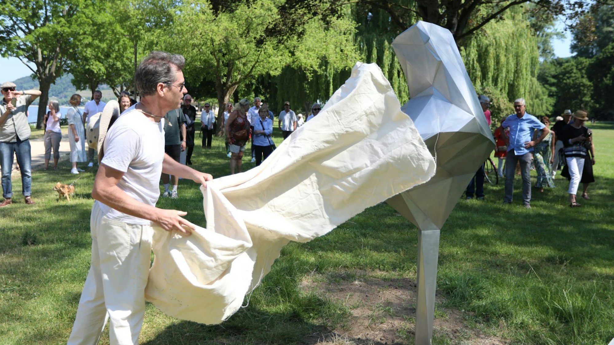 Ein Mann enthüllt ein etwa zwei Meter großes, silbernes Kunstwerk.Im Hintergrund stehende Menschen schauen zu.