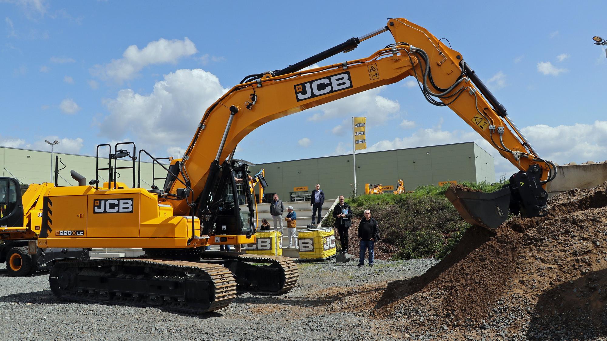 Ein gelber Bagger steht vor einigen Menschen auf einer Baustelle.