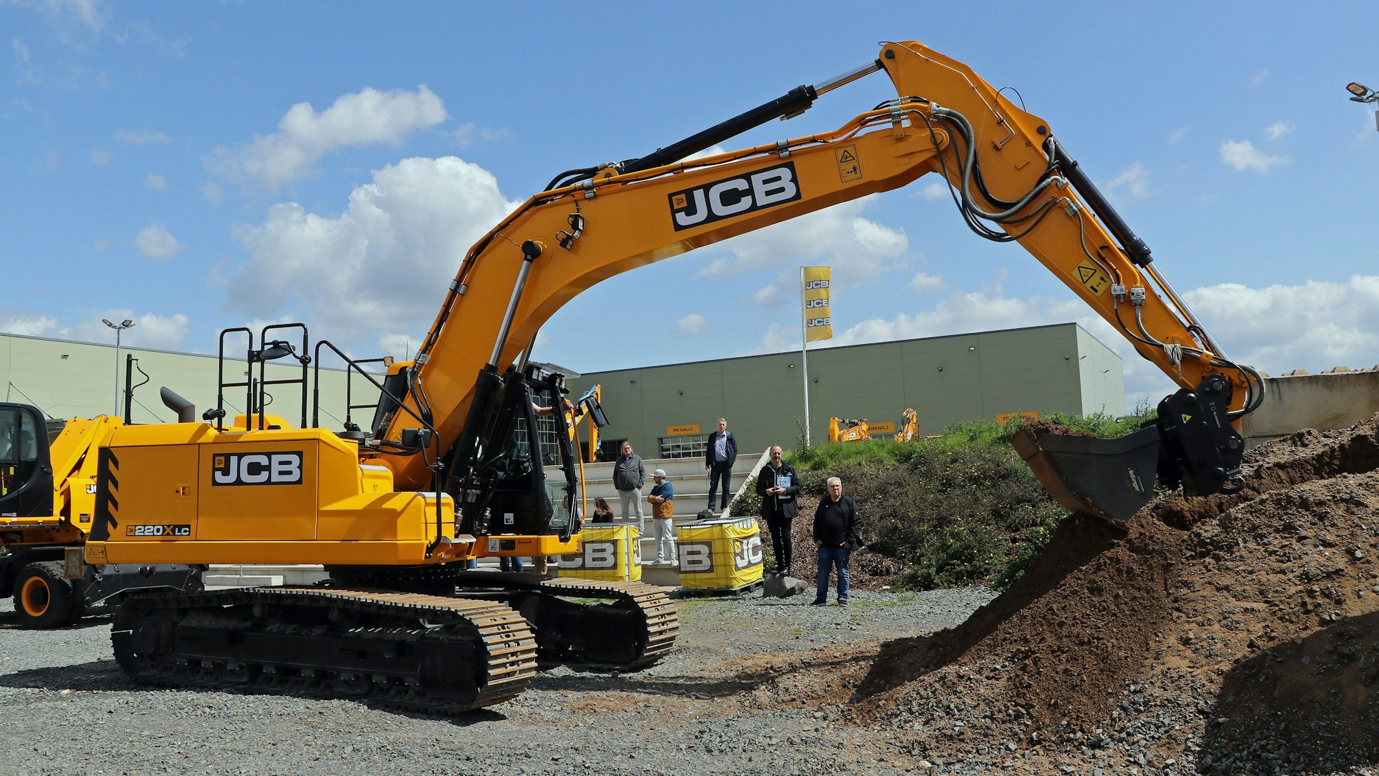 Ein gelber Bagger steht vor einigen Menschen auf einer Baustelle.