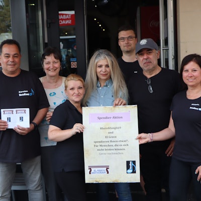 Die Gastronomen des Restaurant El Greco halten ein Hinweisplakat in die Kamera, der besagt, dass Bedürftige bei hnen nun einen Gutschein für Essen oder/und Trinken erhalten.