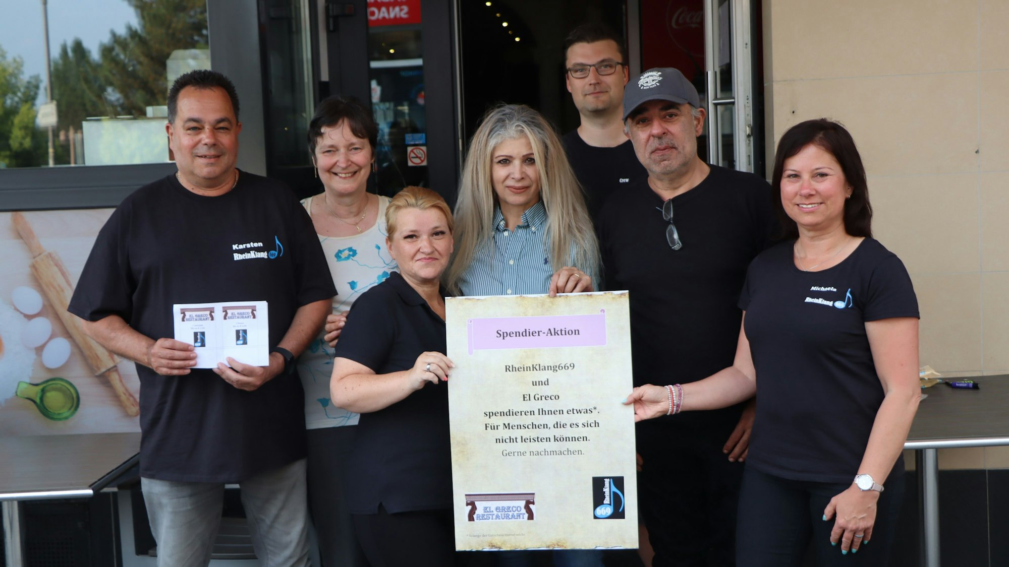 Die Gastronomen des Restaurant El Greco halten ein Hinweisplakat in die Kamera, der besagt, dass Bedürftige bei hnen nun einen Gutschein für Essen oder/und Trinken erhalten.