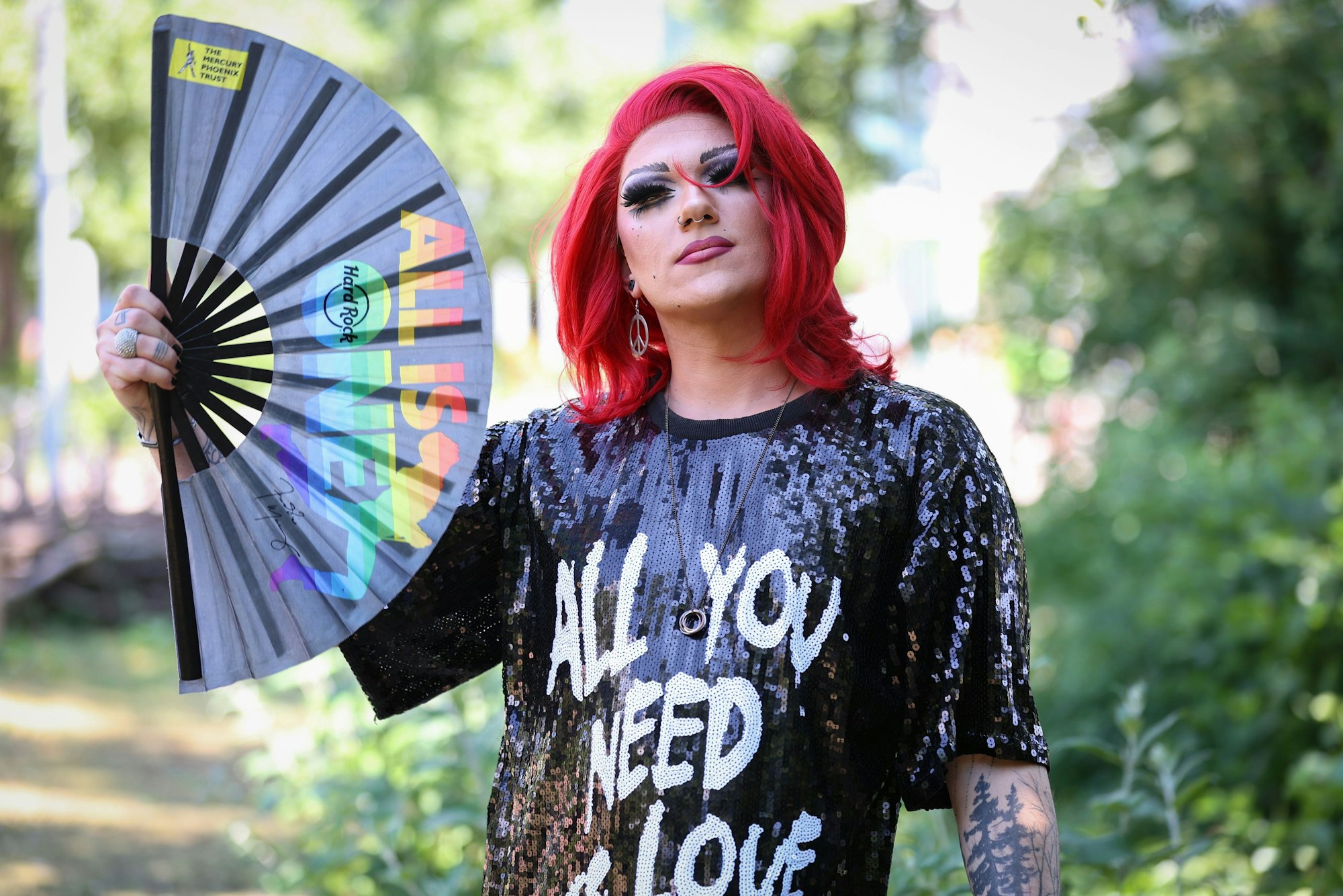 Marcel Kaupp aka Marcella Rockefeller in Drag. Er schaut in die Kamera und hält einen Fächer, auf dem in Regenbogenfarben „All is One“ steht. Auf Marcellas Pallettenkleid steht „All you need is Love“.
