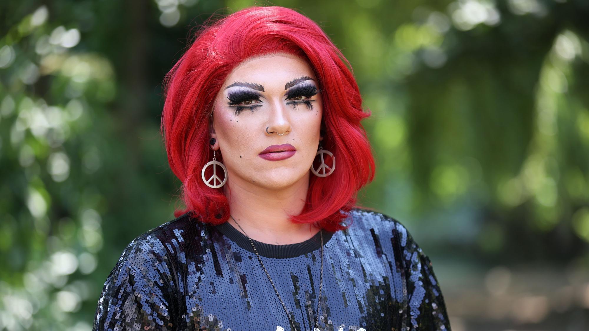 Marcel Kaupp aka Marcella Rockefeller schaut traurig in die Kamera. Die Dragqueen trägt eine rote Perücke und „Peace“-Ohrringe.