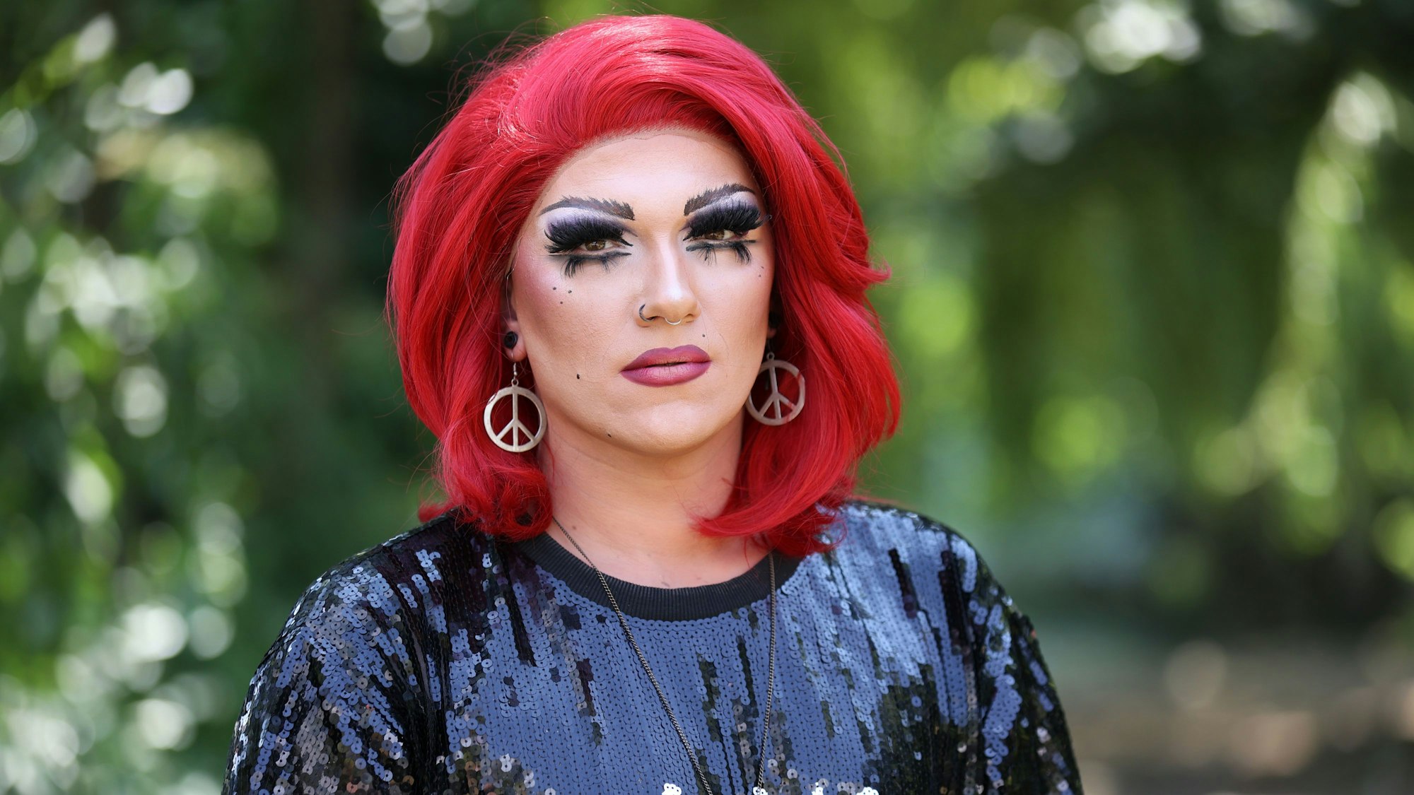 Marcel Kaupp aka Marcella Rockefeller schaut traurig in die Kamera. Die Dragqueen trägt eine rote Perücke und „Peace“-Ohrringe.