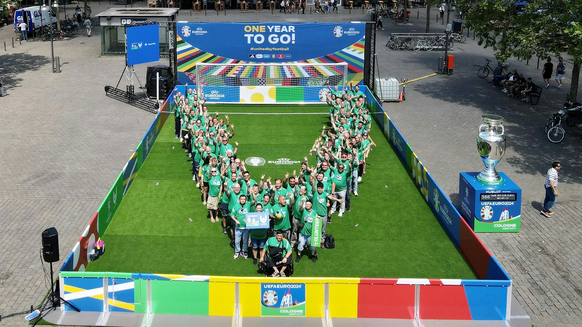 Auf einem Fußballfeld in Miniformat posieren die grün gekleidten Volunteers in Form eines „V“ und formen ein „Victory-Zeichen“ mit den Händen.