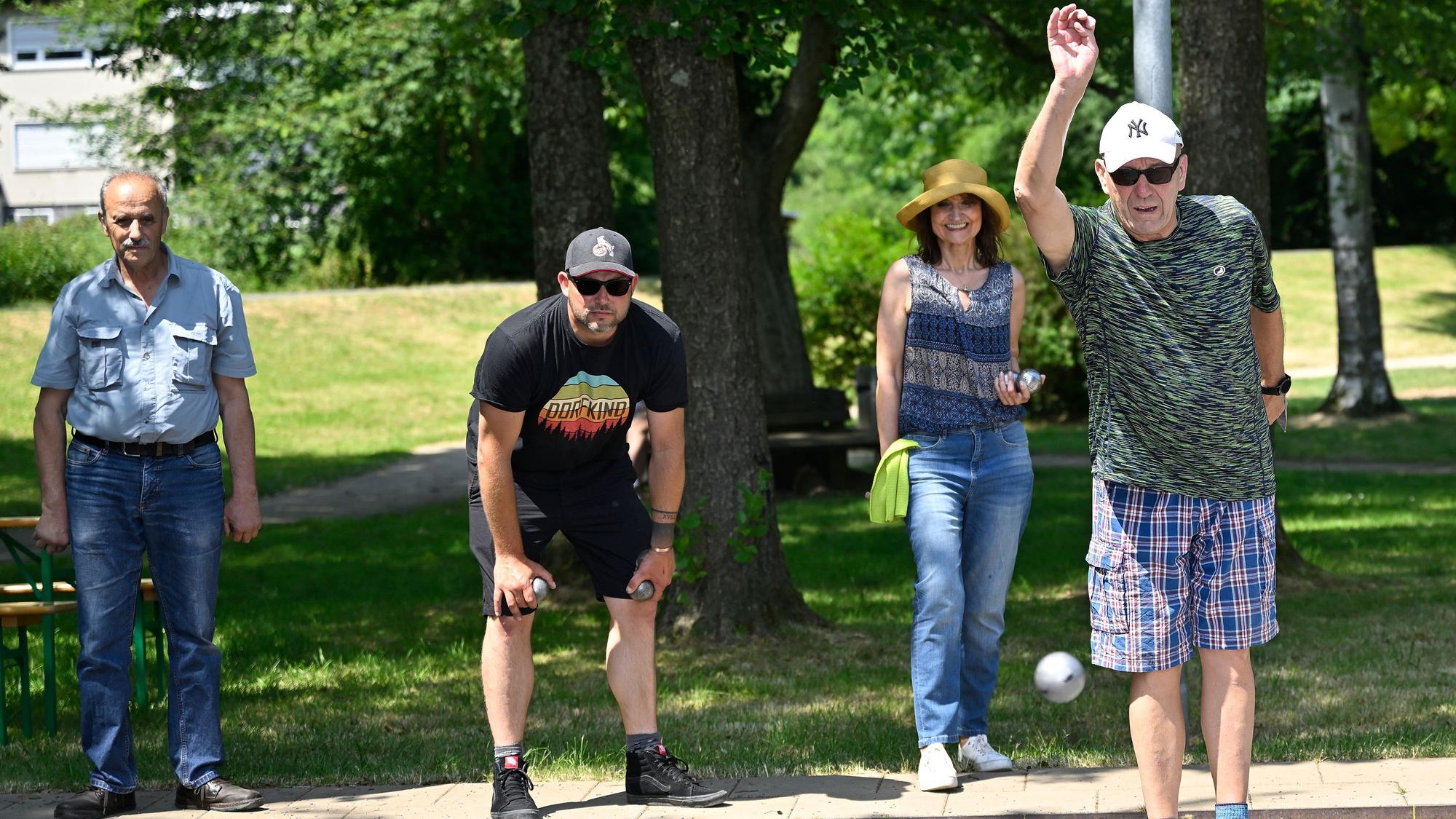 Zwei Männer mit Sonnenbrillen werfen Kugeln in einem Park, im Hintergrund steht eine Frau mit einem Hut und ein Mann.