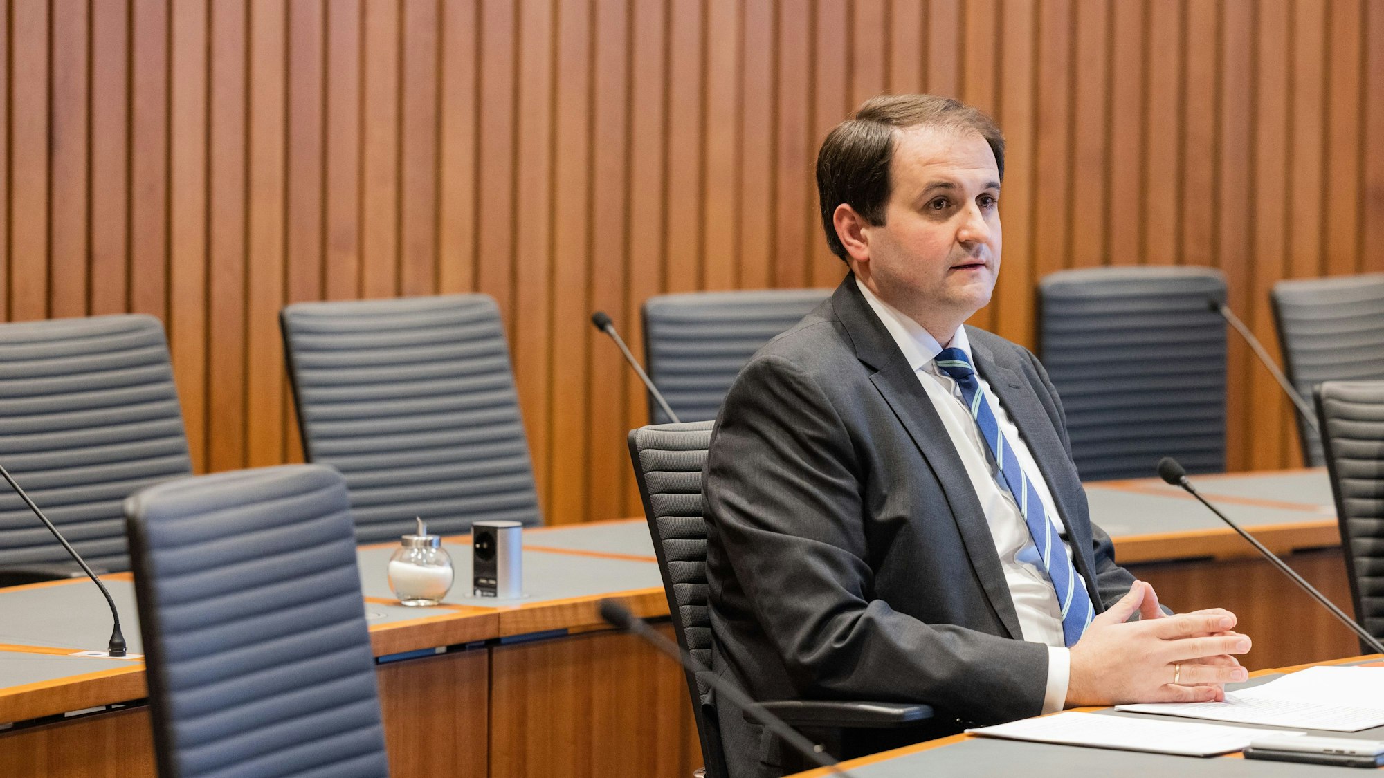 Nathanael Liminski, Chef der Staatskanzlei, sitzt in einem großen, holzvertäfelten Besprechungsraum. Er trägt einen dunkelgrauen Anzug mit blauer Krawatte.