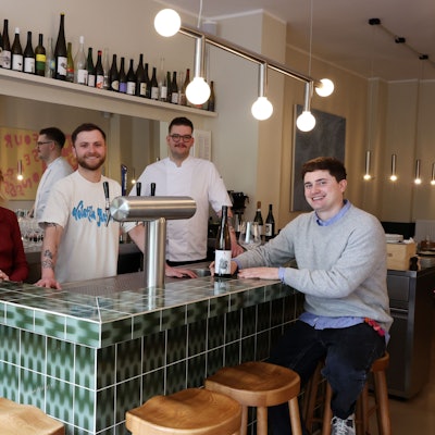 Das vierköpfige Betreiber-Team steht an einer grünen Theke mit Holzhockern.