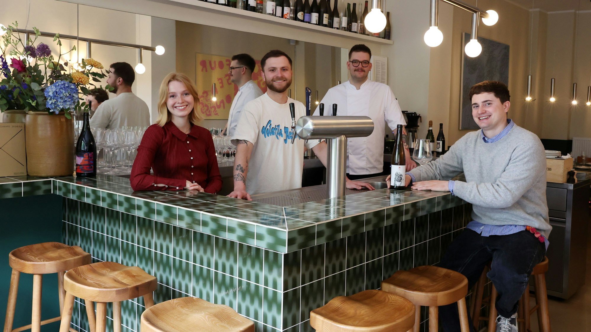Das vierköpfige Betreiber-Team steht an einer grünen Theke mit Holzhockern.