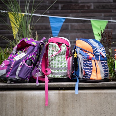 Drei bunte Schulrucksäcke stehen auf einer Bank.