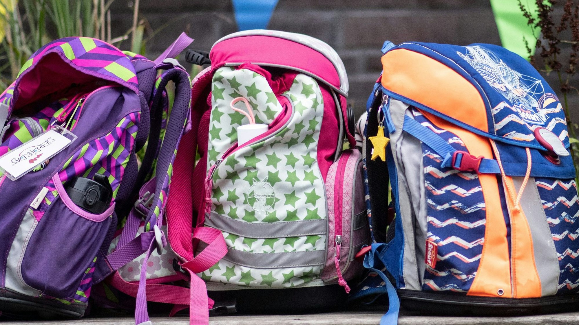 Drei bunte Schulrucksäcke stehen auf einer Bank.