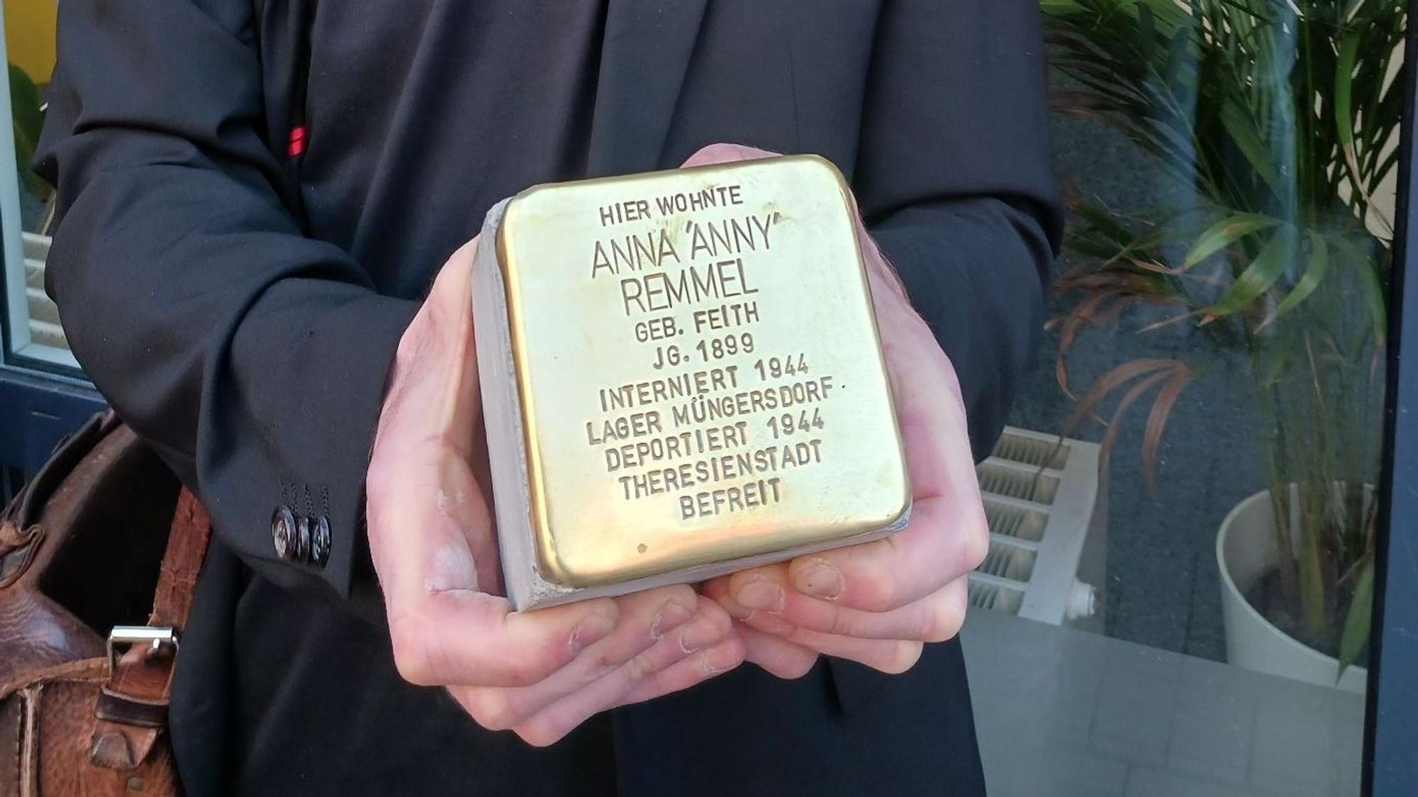 Ein Mann hält einen Pflasterstein, der mit einer goldfarbene Kappe und eingraviertem Text versehen ist.