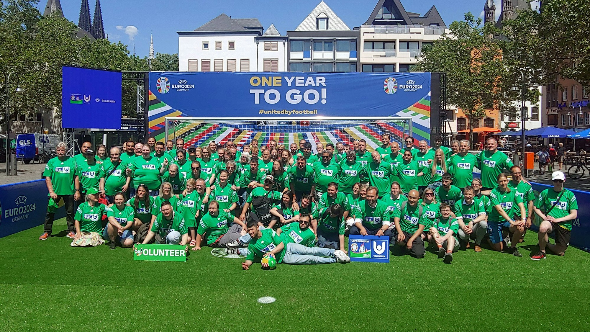 Gruppe von Menschen in grünen T-Shirts auf einem Fußballfeld, die vor einem Tor und einem Schild mit der Aufschrift „One Year To Go!“ stehen