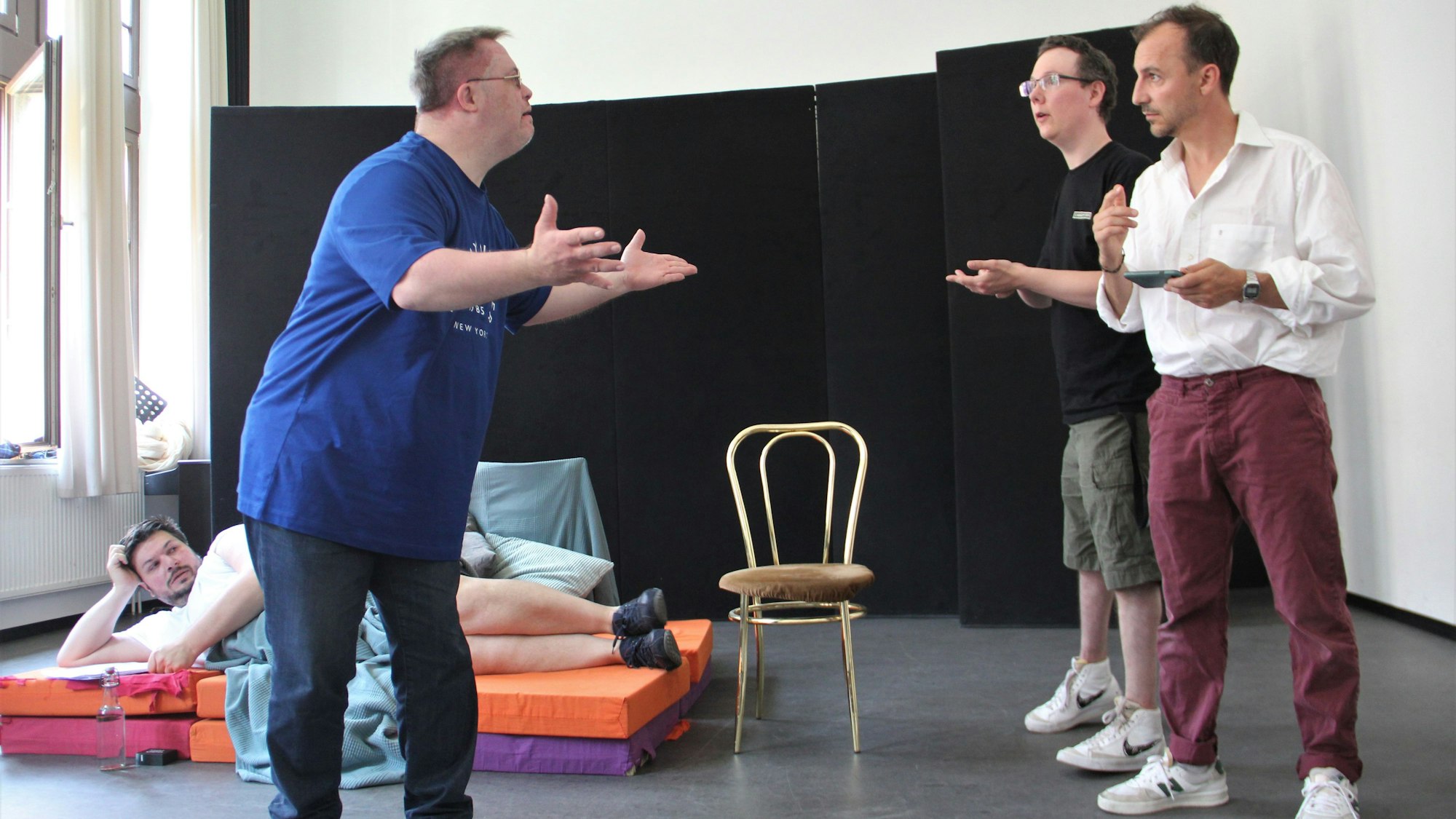 Drei Männer unterhalten sich stehend und gestikulieren, ein vierter Mann liegt im Hintergrund auf einer Matraze