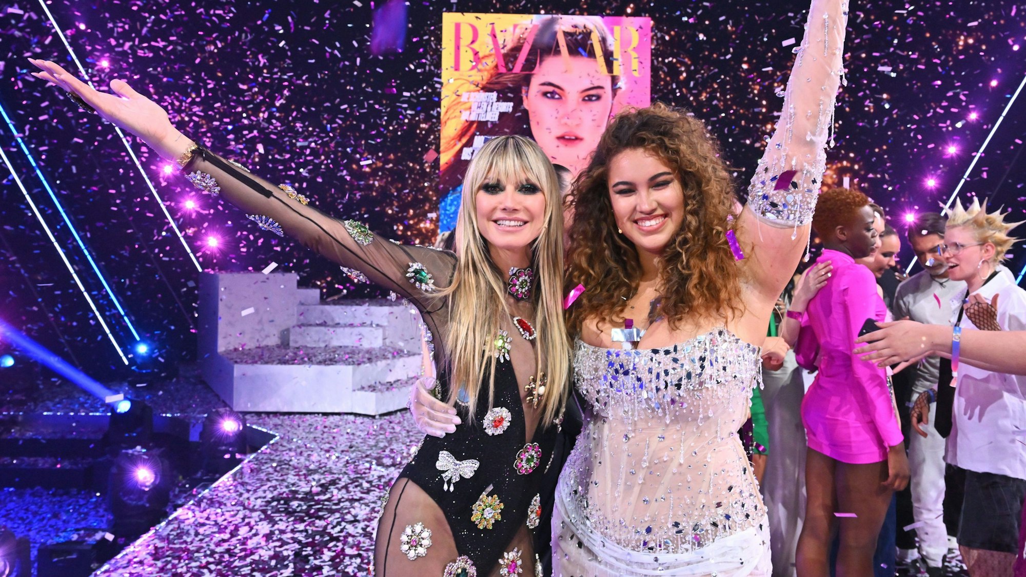 Links steht Heidi Klum neben Vivien (rechts), der Siegerin der 18. Staffel der Casting-Show Germany's Next Topmodel. Es regnet Konfetti von oben, im Hintergrund ist das Cover des Magazins Harpers Bazaar zu sehen, auf dem Vivien abgebildet ist. Heidi und Vivien recken die Arme in die Luft, greifen mit der anderen Hand um die Taille der jeweils anderen Person.