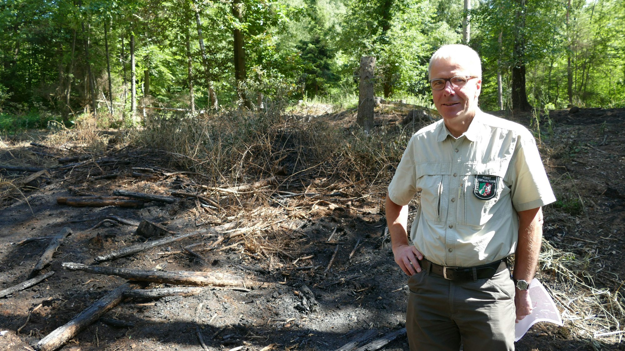Ein älterer Mann, mit dem Aufnäher des Forstamts auf dem Oberhemd, steht auf verkohltem Boden im Wald.