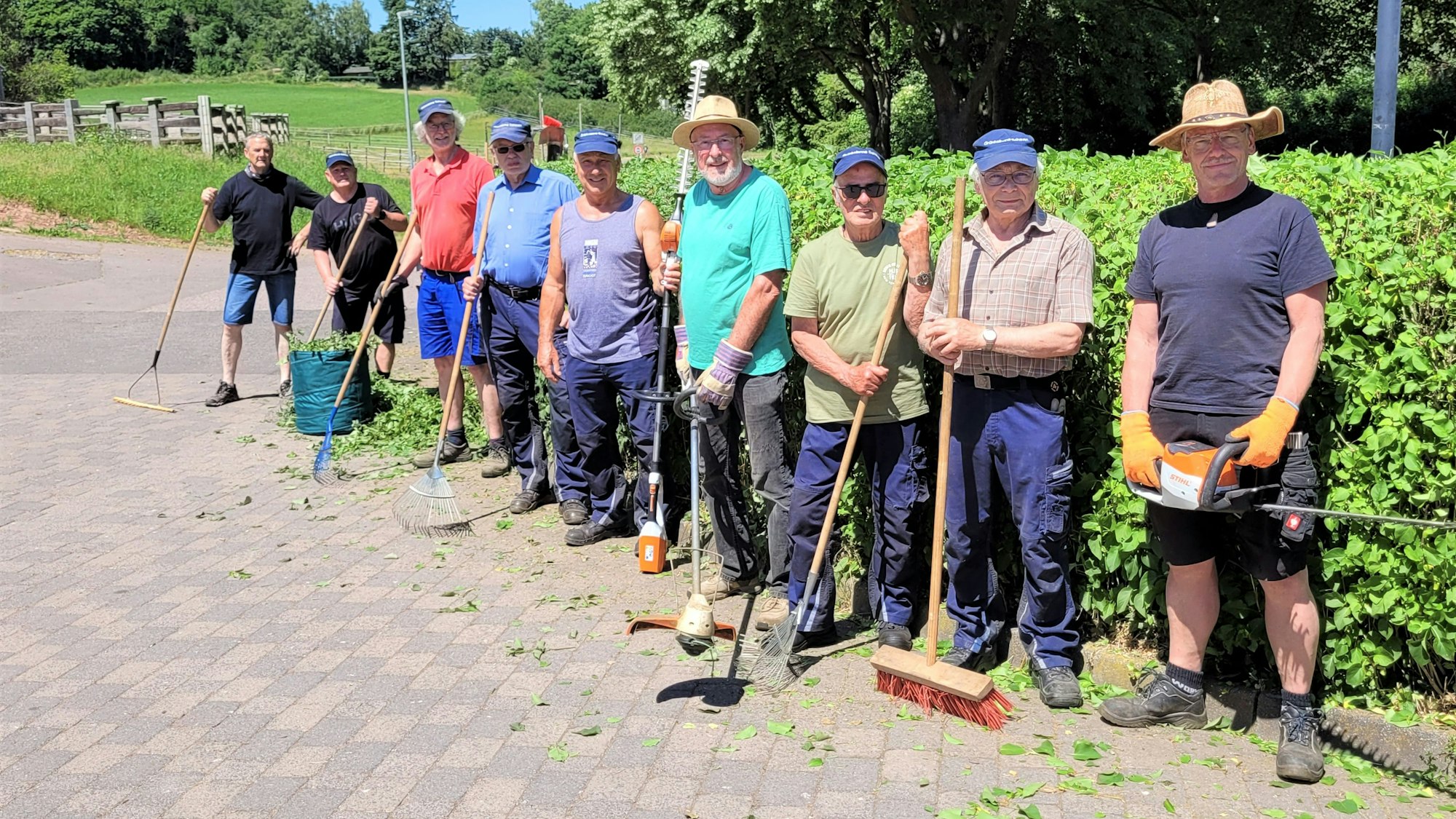 Das Bild zeigt eine Gruppe Männer, die an einer grünen Hecke stehen. Die Männer halten Arbeitsgeräte wie Freischneider oder Besen in den Händen
