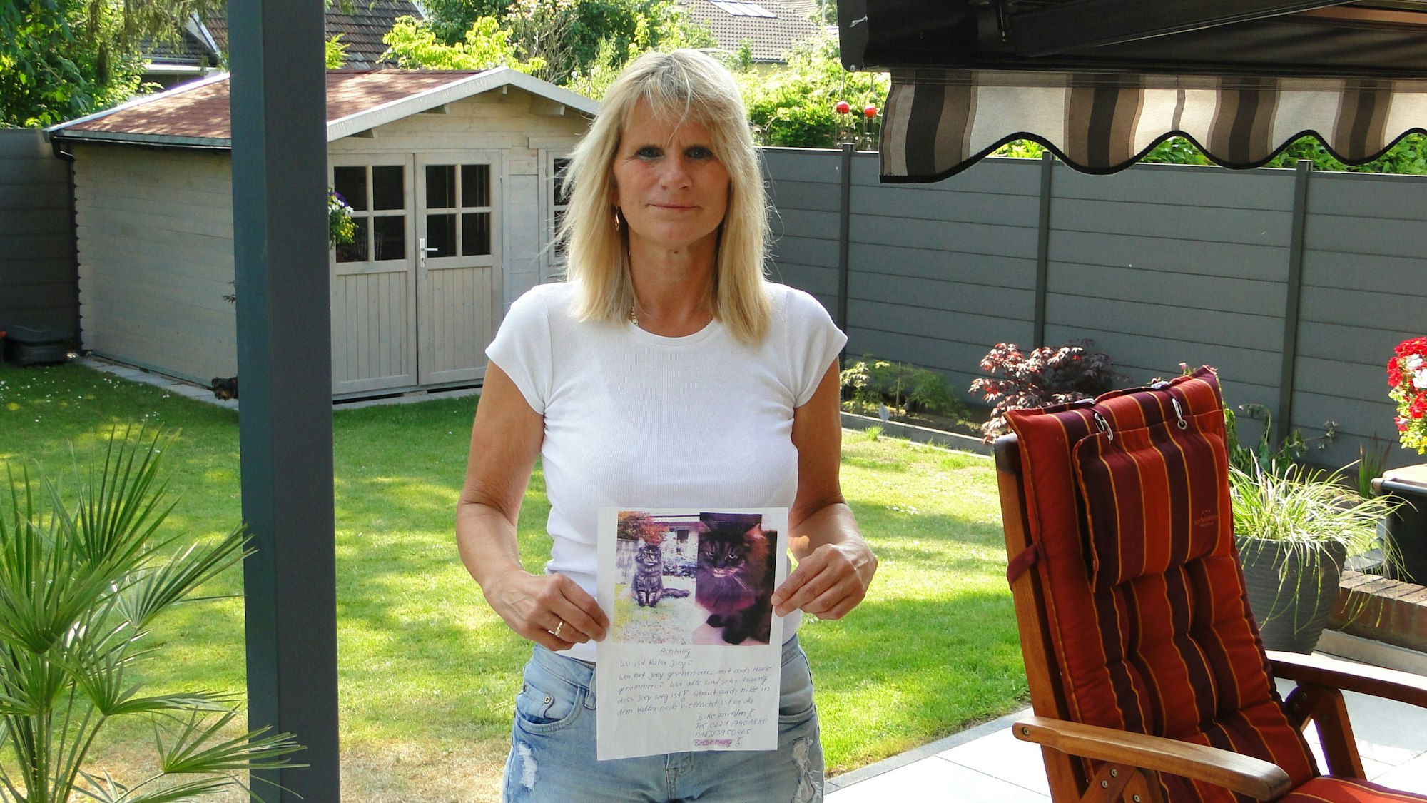 Eine Frau steht auf der Terrasse eines Hauses, ein Rasen und Gartenhaus im Hintergrund. In der Hand hält sie die Suchanzeige für ihren Kater.