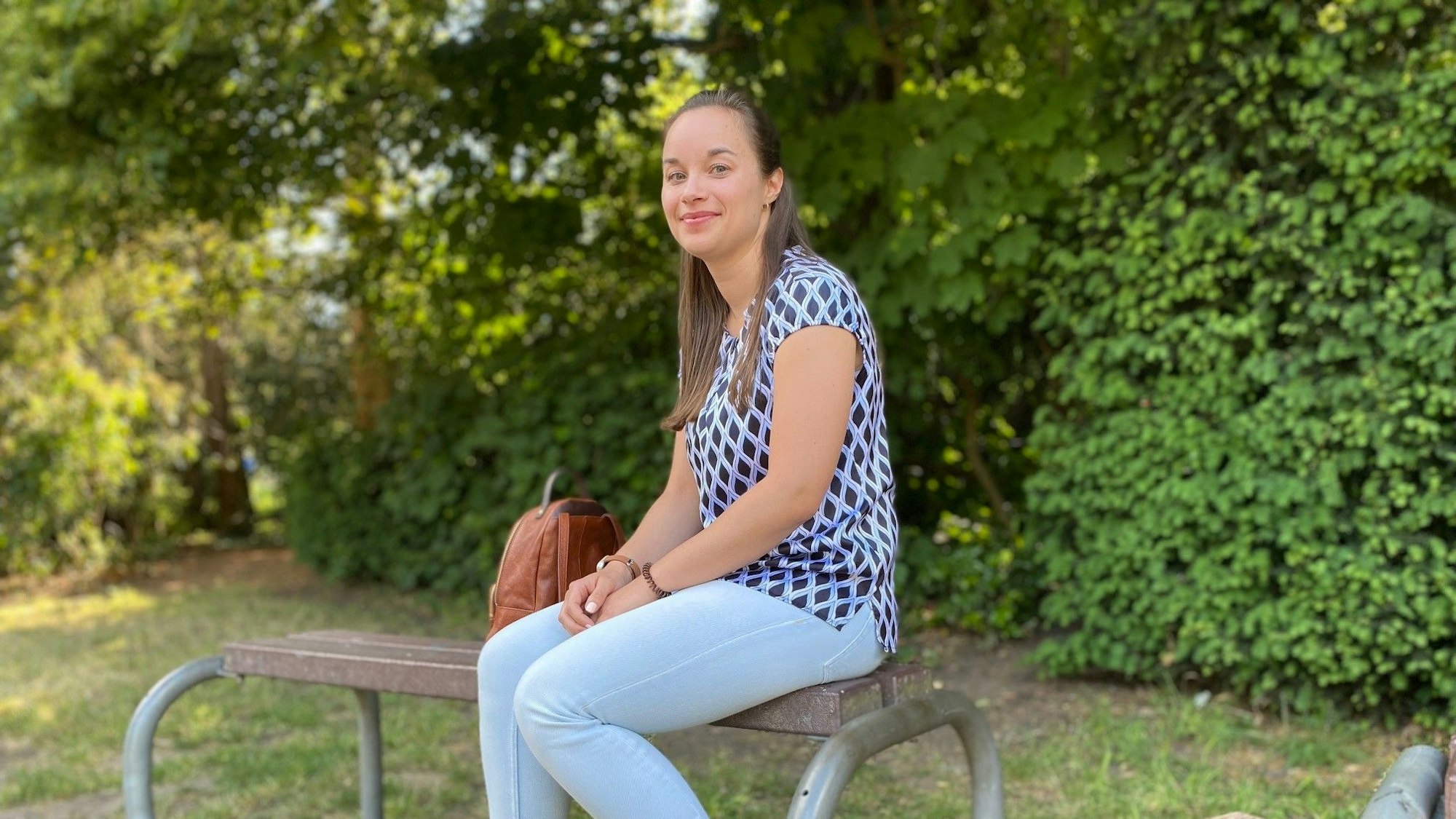 Die junge Frau sitzt auf einer Bank. Sie trägt Jeans und ein gemustertes T-Shirt. Leicht lächelnd blickt sie in die Kamera.