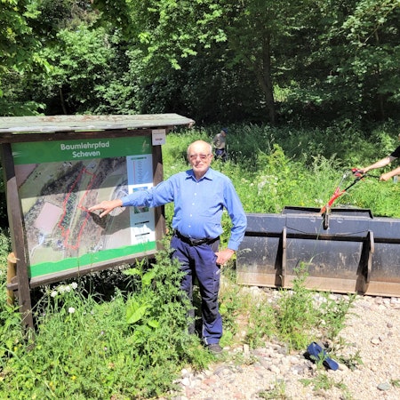 Ein Mann zeigt auf eine Informationstafel, im Hintergrund arbeitet ein weiterer Mann im Grünen.