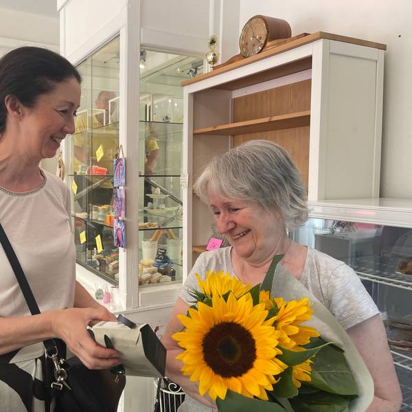 Eine ältere Frau hat einen Strauß Sonnenblumen im Arm, eine dunkelhaarige Frau überreicht ihr noch ein Päckchen. Sie stehen in einem Raum mit Vitrinen und Kaffeekannen.