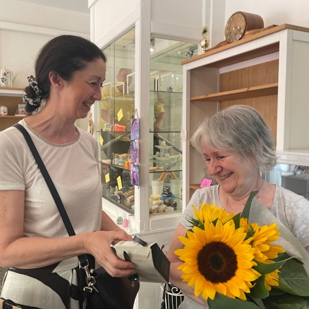 Eine ältere Frau hat einen Strauß Sonnenblumen im Arm, eine dunkelhaarige Frau überreicht ihr noch ein Päckchen. Sie stehen in einem Raum mit Vitrinen und Kaffeekannen.