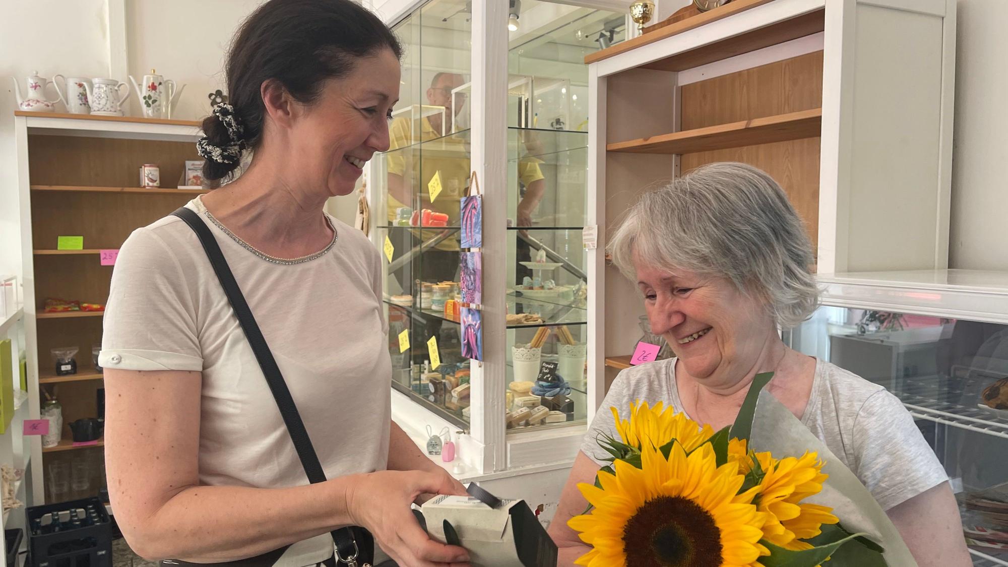 Eine ältere Frau hat einen Strauß Sonnenblumen im Arm, eine dunkelhaarige Frau überreicht ihr noch ein Päckchen. Sie stehen in einem Raum mit Vitrinen und Kaffeekannen.
