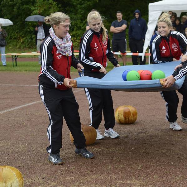 Vier Mädchen in Trainingsanzügen balancieren mehrere Bälle auf einer Turnmatte. Sie befinden sich auf einem Ascheplatz, im Hintergrund finden sich Zuschauer.
