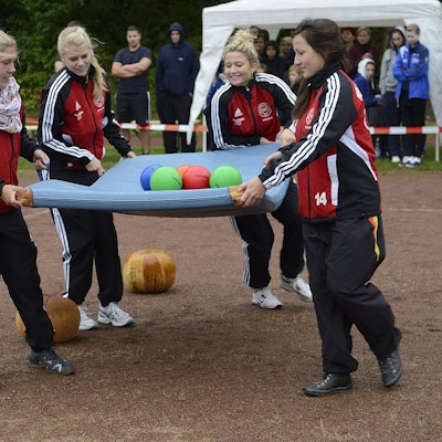 Vier Mädchen in Trainingsanzügen balancieren mehrere Bälle auf einer Turnmatte. Sie befinden sich auf einem Ascheplatz, im Hintergrund finden sich Zuschauer.