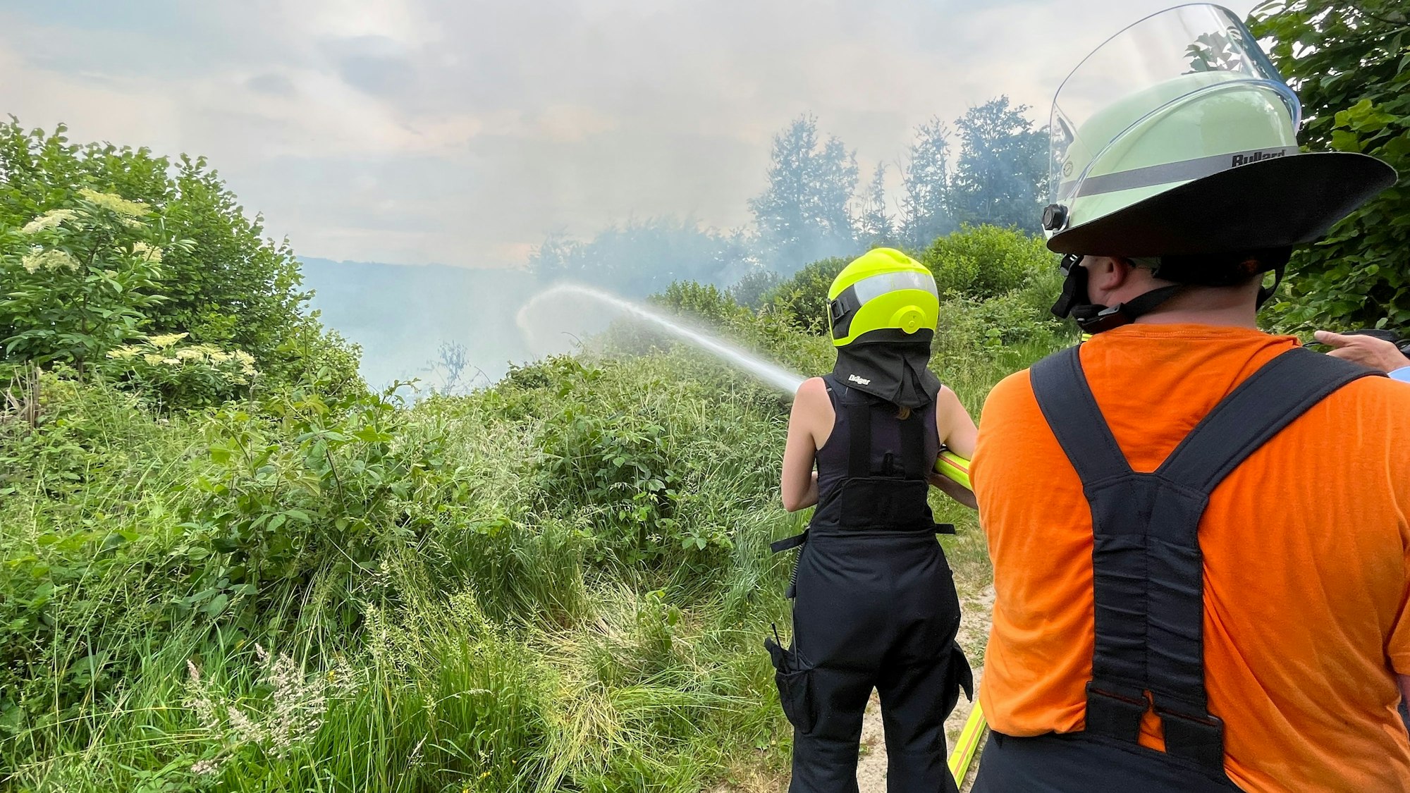Zwei Feuerwehrleute stehen in dichter Vegatation am Waldrand und halten einen dicken Wasserstrahl auf züngelnde Flammen und Rauch.